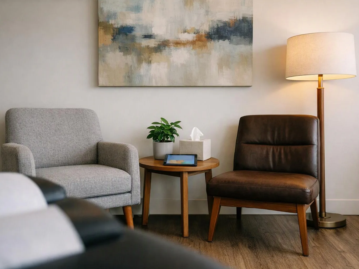 A cozy waiting room has two chairs, a small table, a floor lamp, and abstract wall art