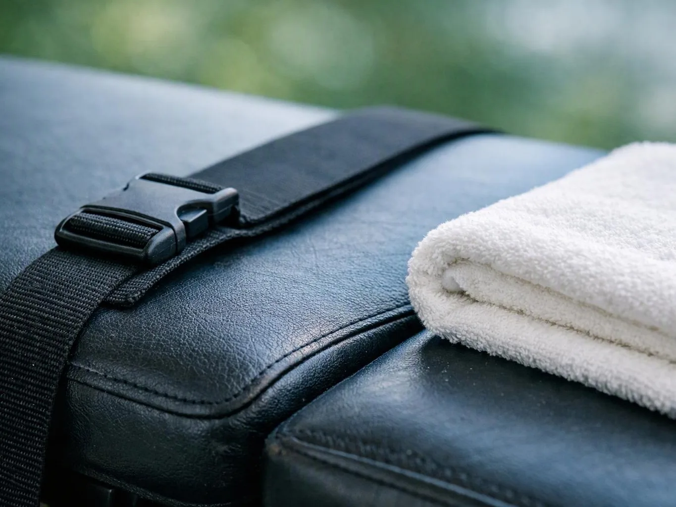 A folded white towel rests on a black leather cushion with a strap and buckle outdoors