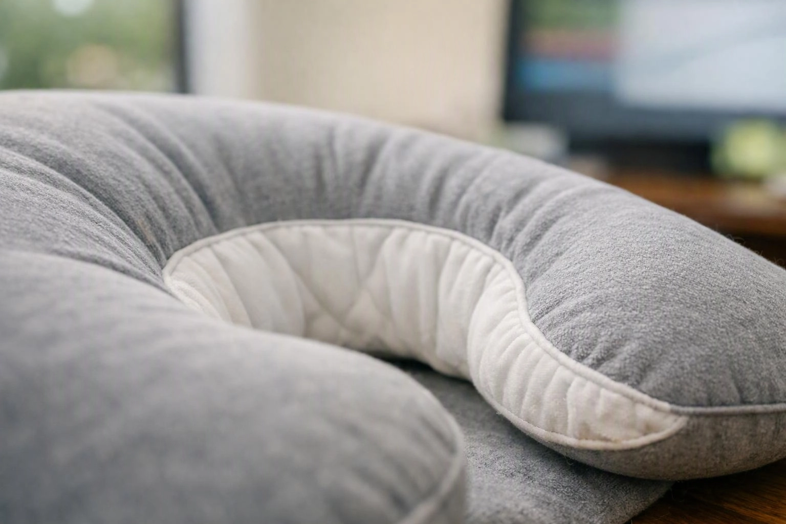 A gray U-shaped cushion rests on a soft surface in a bright room