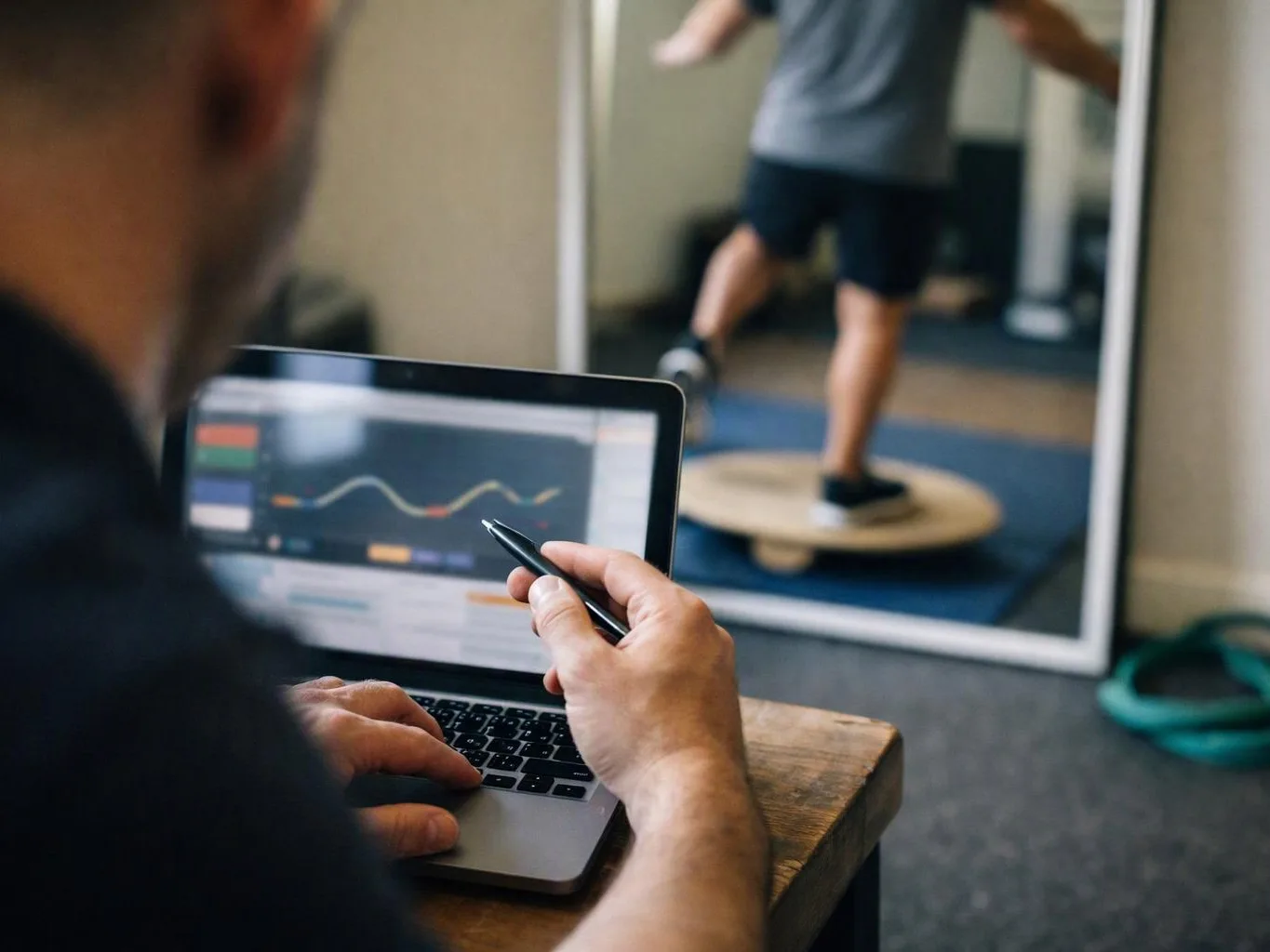 A trainer studies a laptop chart while a person balances on a wobble board in a gym mirror